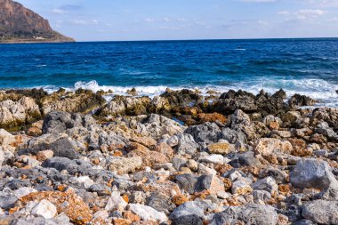 Vivid orange and gray rocks cover the rugged shoreline near Monemvasia, Greece, with foamy blue waves crashing against jagged coastal stones beneath a bright sky.