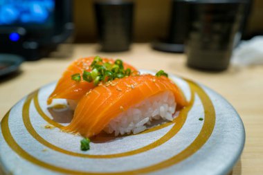 Üzerinde doğranmış yeşil soğan ve susam tohumları bulunan iki parça somon nigiri suşisi altın kıvrımlı gümüş bir tabakta düzenlenir. Parlak balıklar ve narin pirinçler çağdaş bir Japon restoranında hafif bir iç aydınlatma ile vurgulanır..