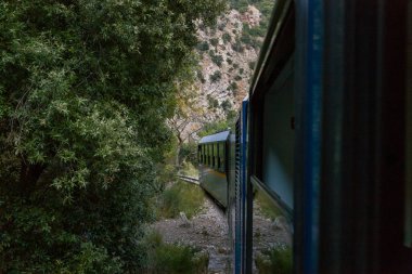 Yunanistan 'ın Kalavrita kenti yakınlarındaki Vouraikos Vadisi' nde yoğun yeşil yapraklar ve engebeli kireçtaşı kayalıklarıyla çevrili dar bir tren yolu kıvrımları çiziyor. Manzara, vahşi doğayı ve bu dramatik dağ yolu boyunca eşsiz yolculuğu yakalar..