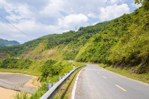 Kuzey Vietnam 'da parçalı bulutlu bir gökyüzünün altında yeşil yamaçlar boyunca metal korkuluklar ve nehir kenarları bulunan sakin bir asfalt yol kıvrımlıdır. Bereketli bitki örtüsü ve parlak gün ışığı yeni ve doğal bir manzara yaratır..