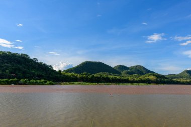 Luang Prabang, Laos yakınlarındaki yemyeşil tepeleri geçen çamurlu Mekong nehrinin geniş manzarası. Açık mavi gökyüzü ve canlı bitki örtüsü seyahat ve doğa kavramları için ideal taze, doğal bir manzara yaratır..