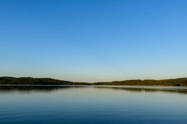Pürüzsüz göl yüzeyi, berrak mavi gökyüzünü ve ağaçlarla kaplı uzak kıyı şeridini yansıtır. Yumuşak bir akşam ışığı, sakin ve sakin bir hava yaratır..