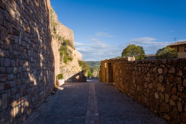 Kırsal taştan duvarlarla çevrili dar kaldırım yolu İspanya 'nın Alquezar kentindeki açık kırlara çıkıyor. Ilık güneş ışığı ve derin gölgeler berrak mavi gökyüzünün altında sakin bir atmosfer yaratır..