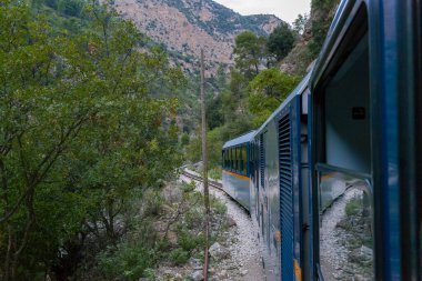 Yunanistan 'ın Kalavrita kenti yakınlarındaki Vouraikos Vadisi' nde yoğun yeşil yapraklar ve engebeli kireçtaşı yamaçlarıyla çevrili mavi dar çaplı bir tren yolu kıvrımları bulunuyor. Sahne dramatik dağ manzarasını ve eşsiz demiryolu yolculuğunu vurgular.