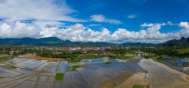 Laos 'taki Vang Vieng' in geniş panoramik manzarası, önplanda pirinç tarlaları ve yeşil dağların eteğinde binalar kümesi. Kabarık beyaz bulutlar ve canlı mavi bir gökyüzü bereketli yağmur mevsimi manzarasını vurgular.