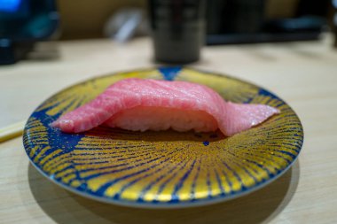 Canlı mavi ve altın desenli tabakta servis edilen tek parça otoro nigiri sushi ve mermer pembe ton balığı. Suşi hafif bir ahşap tezgahta sunuluyor. Hafif aydınlatılmış bir Japon restoranında..