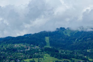 Yoğun bulutlar, Les Portes du Mont Blanc 'ın yamaçlarına yerleşmiş dağınık dağ evleri ve dağlık dağ evleri ile orman tepeleri ve yeşil çayırlar üzerinde sürükleniyor..