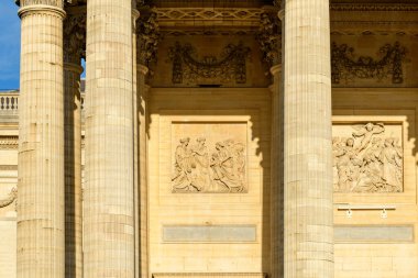 Paris 'teki Pantheon' un güneşli cephesinde neoklasik sütunların ve süslemeli taş yardım panellerinin yakın çekimi, karmaşık oymalar ve mimari dokular sergiliyor..