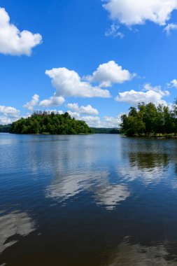 Beyaz kümülüs bulutları ve parlak mavi bir gökyüzü, uzakta ağaçlarla kaplı bir ada ve yeşil kıyı şeridiyle, Settons Gölü 'nün sakin yüzeyinde yansıtılır..
