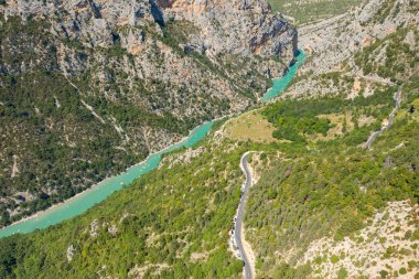 Berrak bir yaz gününde Verdon Vadisi 'ndeki dik kireçtaşı kayalıkları ve yemyeşil çimlerle çevrili turkuaz renkli bir nehre doğru inen dolambaçlı bir dağ yolunun yüksek açılı görüntüsü..