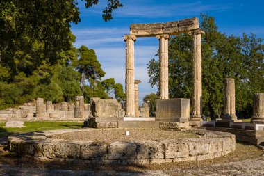 Yunanistan 'ın Olympia kentindeki antik harabeler ve yemyeşil yeşillikler arasında Filipinler' in ayakta duran taş sütunları ve dairesel temelleri yükseliyor. Parlak güneş ışığı bu tarihi arkeolojik alanın klasik mimari biçimlerini ve yıpranmış dokularını vurguluyor.