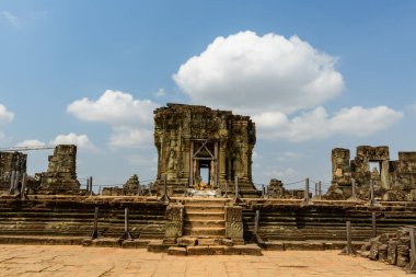 Phnom Bakheng Tapınağı 'nın yıpranmış taş kalıntıları Kamboçya' nın Siem Reap yakınlarındaki bulutlarla dolu parlak bir gökyüzünün altındaki terasta duruyor. Tarihi mekanda ayrıntılı oymalar, kumlu kaldırımlar ve sakin bir arkeolojik dekor yer alıyor..
