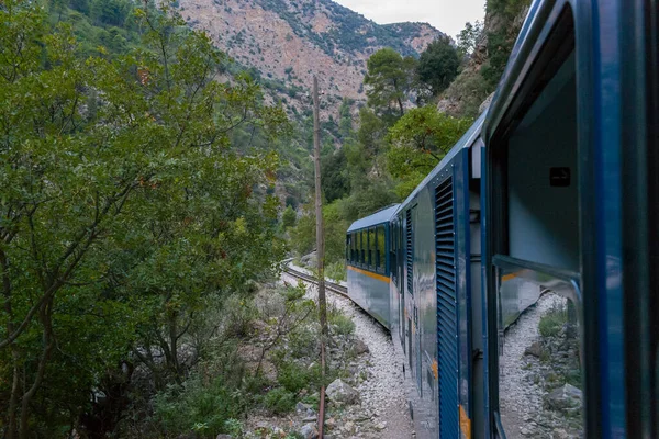 Yunanistan 'ın Kalavrita kenti yakınlarındaki Vouraikos Vadisi' nde yoğun yeşil yapraklar ve engebeli kireçtaşı yamaçlarıyla çevrili mavi dar çaplı bir tren yolu kıvrımları bulunuyor. Sahne dramatik dağ manzarasını ve eşsiz demiryolu yolculuğunu vurgular.