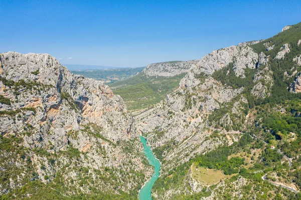 Verdon Vadisi 'ndeki dolambaçlı bir turkuaz nehrin üzerinde yükselen dramatik kireçtaşı uçurumlarının hava perspektifi. Berrak mavi gökyüzünün altında yemyeşil orman ve güneşli tepelerle çevrili..