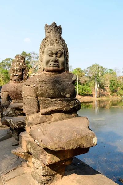 Kamboçya 'daki Siem Reap yakınlarındaki Angkor Thom' a uzanan anıtsal köprüde yıpranmış taştan bir muhafız figürünün yakın çekimi. Anlamlı heykel, tropikal ormanlarla çerçevelenmiş ve gün ışığıyla aydınlatılmış sakin bir hendeğin yanında duruyor..