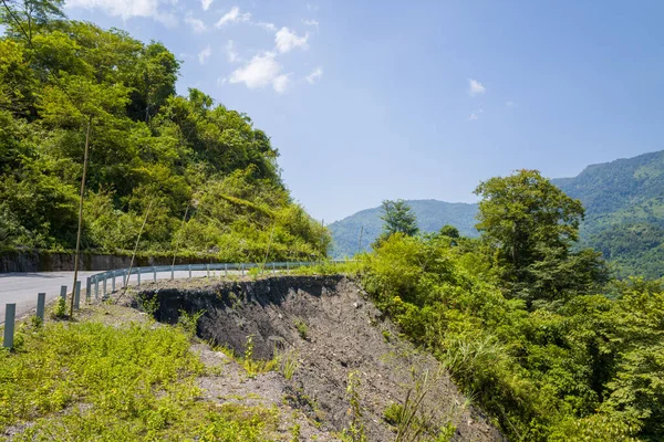 Dik bir heyelan, Lai Chau, Vietnam yakınlarındaki yeşil tepelerde asfalt bir yolun yanında karanlık toprak ve kayalık toprağı ortaya çıkarır. Parlak güneş ışığı açık mavi gökyüzü altında canlı yeşilliği ve dramatik yamacı güçlendirir..