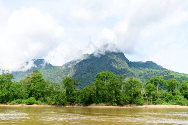 Kuzey Laos 'taki Muang Ngoi yakınlarındaki Nam Ou Nehri boyunca yoğun ormanlık dağların üzerinde beyaz bulutlar yüzer. Sahnede canlı yeşil yapraklar, çamurlu nehir suyu ve parlak, hafif bulutlu bir gökyüzü yer alıyor..