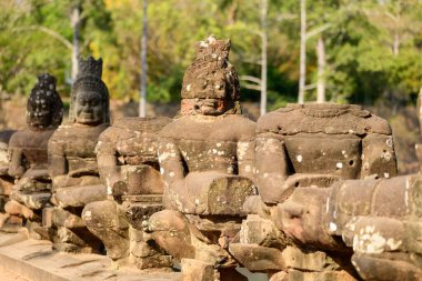 Bir dizi antik taş muhafız heykeli, bir sürü kayıp kafa ve kol, Kamboçya 'daki Siem Reap yakınlarındaki Angkor Thom' un girişindeki köprüyü çiziyor. Zaman aşımına uğramış Khmer heykelleri, yüzyılların altını çizerek günışığıyla aydınlatılmış tropikal ağaçların bir zeminine kurulur.