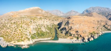 Kurak dağlar ve engebeli uçurumlarla çevrili kumlu sahil ve turkuaz Akdeniz 'le buluşmak için gür palmiye kaplı bir vadiden akan Preveli nehrinin havadan görünüşü berrak mavi bir gökyüzü altında..