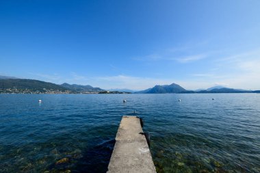 Maggiore Gölü 'nün berrak mavi sularına uzanan taştan bir iskele, geniş ve bulutsuz bir gökyüzünün altında dağınık şamandıralar ve uzak dağ siluetleri ile. Sakin sahne, kuzeydeki şeffaf kıyı şeridi ve panoramik alp zeminini yakalıyor.