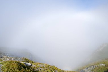 Yoğun sis örtüleri, kayalık alp arazisi, dağ manzarasını gizliyor ve yumuşak gün ışığı altında gizemli, ruhani bir atmosfer yaratıyor..