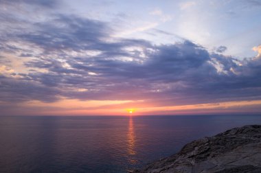 Sardinya 'nın Stintino yakınlarındaki engebeli kayalıkların üzerinde dokulu bulutlar sürüklenirken, gün batımının canlı renkleri sakin Akdeniz sularında yansıyor. Sahne, yumuşak, parlayan ışıkla huzurlu ve atmosferik bir hava yayıyor..