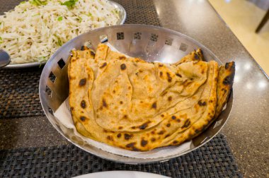 Restoran masasında delinmiş metal bir sepetin içinde servis edilen altın sarısı tandoori paratha ekmeğine yakın plan, arka planda bir tabak kokulu basmati pilavı. Sahnede sıcak bir ışık ve sıradan bir yemek atmosferi var..