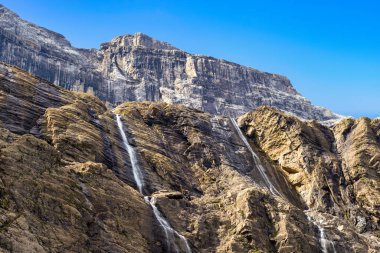 Cirque de Gavarnie 'deki katmanlı kaya kayalıkları parlak güneş ışığıyla aydınlatılır ve birkaç dar şelale dalgalı mavi gökyüzünün altında engebeli kayalardan aşağı akar. Çizgili dokular ve toprak renkler dramatik dağ manzarasını vurgular..