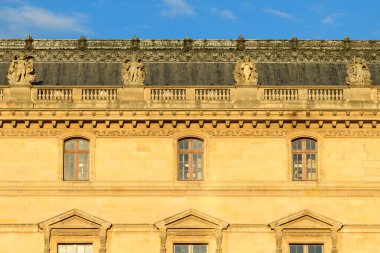 Paris, Fransa 'daki Louvre Sarayı' nın üst çatısında sıcak akşam güneşiyle aydınlatılan taştan heykeller ve balüstreler var. Tarihi cephede simetrik pencereler, karmaşık kornişler ve ayrıntılı rönesans yer alıyor.