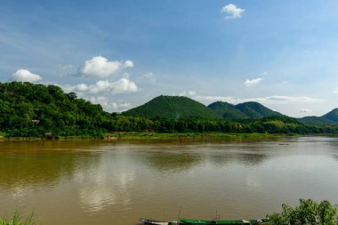 Luang Prabang, Laos yakınlarındaki parlak mavi gökyüzünün altındaki yemyeşil tepelerden akan Mekong nehrinin geniş manzarası. Huzurlu manzara, nehir kıyısı boyunca yumuşak su yansımaları, dağınık bulutlar ve yoğun tropikal bitki örtüsü içerir..