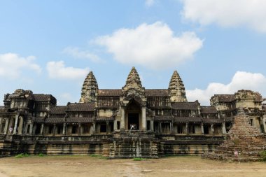 Angkor Vat, Kamboçya 'daki ana kumtaşı cephesinin ön perspektifi, ikonik Khmer kuleleri ve dağınık bulutlu parlak mavi gökyüzünün altında dengeli mimari çizgiler sergiliyor. Tarihi yapı geniş taş basamakların ve kuru