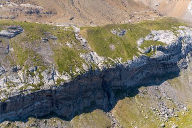 Gavarnie Gedre, Pyrenees yakınlarındaki dağlarda güneşli yeşil bir platonun altına çarpıcı bir kireçtaşı uçurumu düşüyor. Sahne sert kaya dokularını, seyrek bitki örtüsünü ve doğal toprak tonlarını ön plana çıkarıyor..