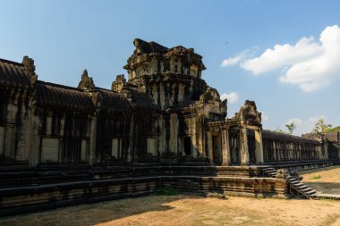 Angkor Vat 'ın giriş köşkünün yıpranmış kumtaşı cephesi öğleden sonra güneş ışığıyla aydınlanır ve karmaşık oymalar ve geometrik çizgiler ortaya çıkar. Kuru çimenler ve dağınık bulutlu açık mavi bir gökyüzü ikonik Khmer mimarisini çerçeveler..