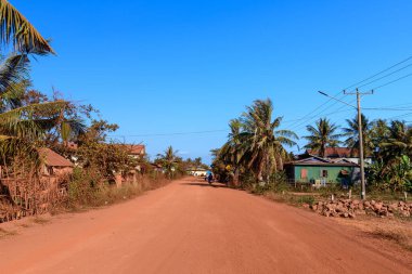 Kamboçya 'nın Kamboçya kentinin kırsal kesimindeki basit evler ve palmiye ağaçları arasında geniş asfaltsız bir toprak yolu bulunur. Sahne berrak güneş ışığıyla yıkanmış, tropikal bitki örtüsünü, kırsal çitleri ve sakin atmosferi vurguluyor.