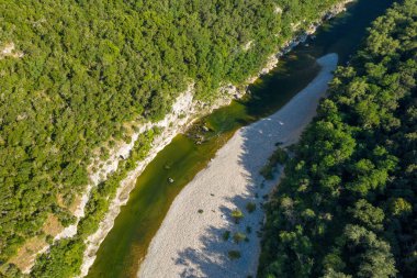 Yoğun yeşil orman ve Ardeche Boğazı 'ndaki kayalık kireçtaşı bankalarla çevrili dolambaçlı bir nehrin yüksek açılı manzarası. Güneş ışığı çakıl taşı plajında taze gölgeler bırakır ve doğal dokuları ön plana çıkarır..