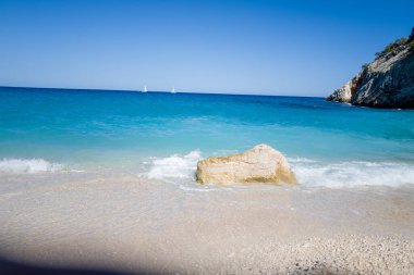 Sardunya 'nın Cala Goloritze kentinin kumlu çakıl taşlı sahilinde hafif turkuaz dalgalar tur atarken, tek bir soluk kaya su kenarında duruyor. Uzaktaki yelkenliler ve engebeli kayalıklar berrak mavi gökyüzünün altındaki sakin sahneyi tamamlıyor..
