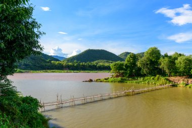Luang Prabang, Laos 'taki yemyeşil tepelerin yakınındaki çamurlu Mekong nehri boyunca kırsal bir bambu köprü uzanır. Nehir kenarındaki yoğun bitki örtüsü ve canlı bir mavi gökyüzü seyahat ve doğa temaları için ideal canlı bir doğal manzara yaratır..