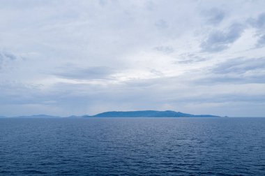 Uçsuz bucaksız mavi su bulutlu bir gökyüzünün altındaki Sardunya 'nın alçak ve puslu hatlarına doğru uzanıyor. Sakin deniz manzarası, sakin ve açık bir alan hissi uyandırıyor..
