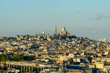 Sacre-Cur bazilikası Montmartre 'ın yoğun dolu çatılarından yükselir, açık mavi gökyüzünün altında sıcak akşam güneşiyle yıkanır. Yükseltilmiş manzara, katmanlı şehir manzarası ve ikonik Paris mimarisini vurguluyor.