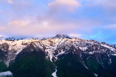 Ilık sabah ışığı, Mont Blanc 'ın engebeli, karla kaplı tepelerini aydınlatır. Altında yeşil ormanlar ve tepesinde pastel bulutlar vardır..