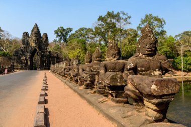 Kamboçya 'nın Siem Reap yakınlarındaki Angkor Thom' un anıtsal güney kapısına uzanan köprü, devasa taş muhafız figürlerinden oluşuyor. Tarihi Khmer heykelleri tropikal ormanlara ve berrak gün ışığında sakin bir hendeğe karşı sergilenir.