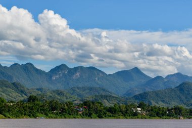Mekong Nehri 'nin yemyeşil tepeleri ve yoğun ormanı geçtikten sonra Luang Prabang, Laos yakınlarındaki dramatik mavi dağ sırtlarıyla sakin bir görüntüsü. Kabarık kümülüs bulutları canlı bir gökyüzünde sürükleniyor ve genişleyen doğaya derinlik katıyor.