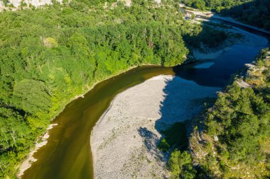 Dönek bir nehir, Gorges de Ardeche 'de sık yeşil ormanlarla çevrili geniş bir çakıl barının etrafında akar. Güneş ışığı, kayaların ve yaprakların dokusunu aydınlatan keskin gölgeler bırakır..