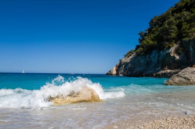 Sardinya, Cala Goloritze 'de turkuvaz suyu ve sarp kayalıklarla kaplı bir çakıl taşı plajında köpüklü bir dalga kırılır. Açık mavi gökyüzü ve canlı deniz dinamik, yaz atmosferi yaratır..