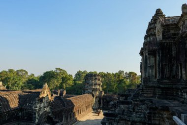 Angkor Vat 'ın zirvesine yükseltilmiş perspektif, yoğun tropikal ormanlara ve geniş mavi gökyüzüne karşı kurulmuş yıpranmış taş galerileri ve süslü çatı hatlarını ortaya çıkarıyor. Antik Khmer mimarisi sıcak güneş ışığı ve karmaşık detaylarla vurgulanır..