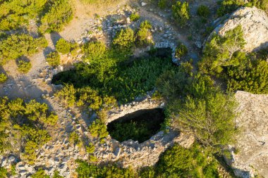 Yıpranmış bir taş kalıntısının havadan görünüşü yoğun yeşil bitki örtüsüyle dolu, kayalık araziler ve sıcak akşam güneşi altında Opoul Şatosu yakınlarındaki dağınık çalılarla çevrili..