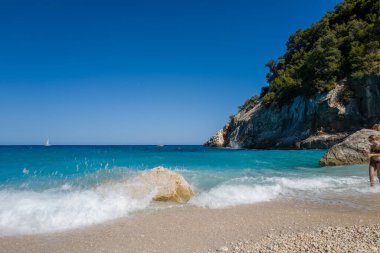 Sardinya 'nın Cala Goloritze kentindeki körfezi çevreleyen çarpıcı kireçtaşı kayalıkları ve yemyeşil yeşil kayalıklarıyla çakıllı bir plajda güneşli kayalara vuran parlak turkuaz dalgalar. Açık mavi gökyüzü ve canlı su ferahlatıcı bir yaz havası uyandırıyor..