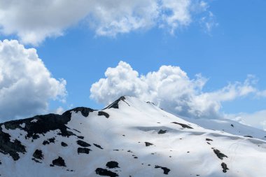 Aiguillette des Houches yumuşak bir dağ zirvesi sergiliyor eriyen kar parçalarıyla ve parlak gökyüzünün altında ortaya çıkan koyu renkli kayalar tüylü kümülüs bulutlarıyla dolu..