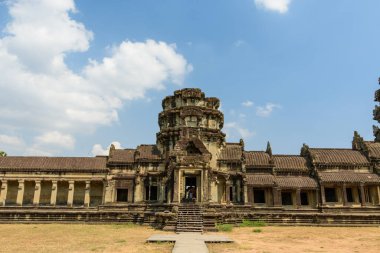 Angkor Wat 'ın merkez kumtaşı giriş pavyonu dağınık bulutlarla dolu açık mavi bir gökyüzünün altında güneş ışığı altında duruyor. Yıpranmış taş işçiliği, simetrik sütunlar ve geometrik çizgiler bu ikonik Khmer tapınağı kompleksinin ihtişamını vurguluyor..