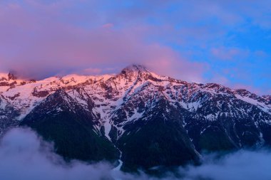 Canlı pembe ve mor alpenglow, Mont Blanc 'ın karla kaplı zirvesini aydınlatıyor. Aşağıda sakin akşam atmosferinde dramatik bulutlar ve karanlık orman yamaçları var..